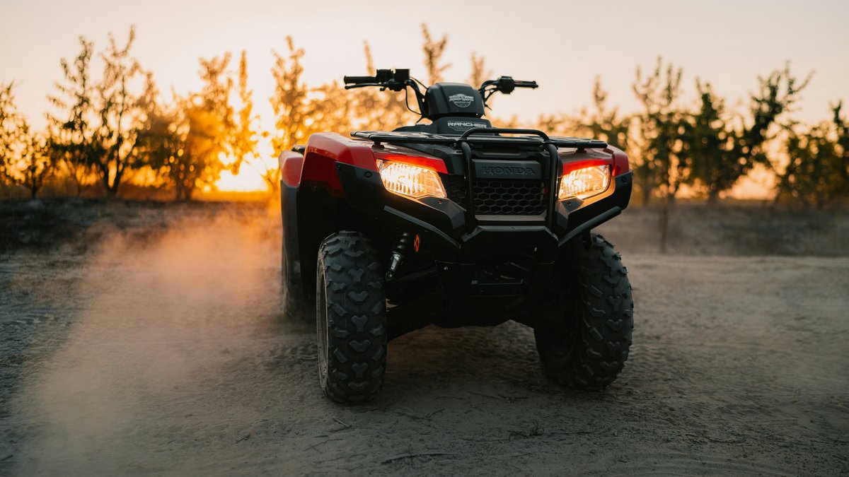 Red Honda utility ATV kicks up dust on a trail at sunset. Bright LED headlights, knobby off-road tires.